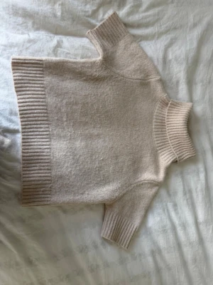 Beige stickad tröja med polokrage - Mysig beige stickad tröja med korta ärmar och bred polokrage. Tröjan har ribbstickade detaljer vid hals, ärmslut och nederkant. Perfekt för lager-på-lager och enkel att matcha med olika outfits.