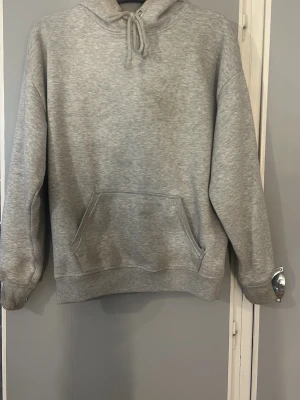 Grå basic hoodie med huva - En enkel grå hoodie med huva och dragsko, samt en stor känguruficka framtill. Tillverkad i mjukt sweatshirtmaterial som är perfekt för chill dagar. Klassisk passform och ribbade muddar vid ärmar och nederkant. Den har lite fläckar