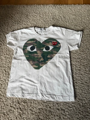 Vit Comme des Garçons PLAY t-shirt - Vit t-shirt från Comme des Garçons PLAY med stort hjärta i camouflage-mönster och ett litet rött broderat hjärta med ögon på bröstet. Klassisk rund hals och korta ärmar. Perfekt statement-plagg för din garderob.
