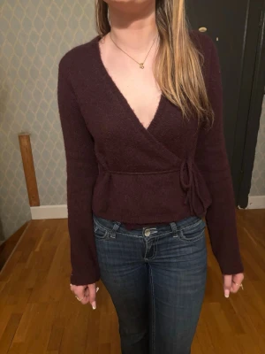 Vinröd omlottkofta med knytband - Mysig vinröd omlottkofta med långa ärmar och knytband i midjan. Koftan har en djup v-ringning och är stickad i ett mjukt material som känns skönt mot huden. Perfekt för lager-på-lager och en chill look.