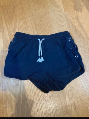 Svarta shorts med snörning i sidan - Snygga svarta shorts med elastisk midja och vit dragsko framtill. På sidorna finns cool snörningsdetalj med metallöljetter som ger en sportig vibe. Tillverkade i mjukt bomullstyg, perfekta för sommardagar eller träning. Barnstorlek 146/152 men passar även xxs.