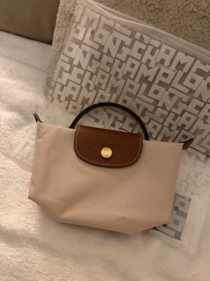 Beige Longchamp handväska med läderdetaljer - Snygg beige handväska från Longchamp med klassisk brun läderdetalj och guldfärgad knapp framtill. Väskan har en stilren, kompakt design med korta handtag i brunt läder. Perfekt för dig som gillar enkel och tidlös stil. Köpt hos longchamp.com. Finns en liten prick på väskan man syns knappt. Finns kvitto på mail.