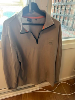 Beige halfzip tröja från BOSS - Snygg beige halfzip tröja från BOSS med svart dragkedja och orange detaljer på insidan av kragen. Tröjan har en hög krage och BOSS-logga på bröstet. Perfekt för en avslappnad men stilren look.