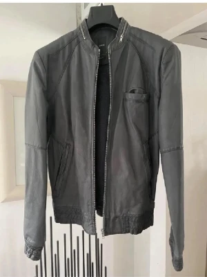 Grå skinnjacka med dragkedja - Snygg skinnjacka med rak passform och dragkedja framtill. Jackan har ståkrage, två snedställda fickor och en bröstficka. Tidlös design som passar till det mesta och ger en cool look.