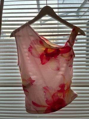 Blommig oneshoulder topp i rosa toner - Säljer en snygg oneshoulder topp med stort blommigt mönster i rosa, rött och gult. Toppen har en asymmetrisk axel och är figurnära med stretchigt material. Perfekt för dig som vill sticka ut lite extra!