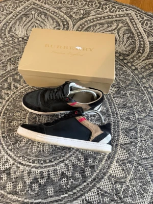 Burberry sneakers med rutigt mönster - Snygga sneakers från Burberry i svart läder och textil med klassiskt rutigt mönster i beige, svart och rött på sidorna. Lågt skaft och vit sula ger en clean look. Perfekta för dig som vill ha något unikt och stilrent från ett lyxigt märke. Små i storleken.