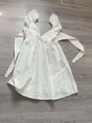 Vit kort klänning med knytband - Söt vit kortklänning med v-ringad front och långa knytband vid axlarna. Klänningen har en luftig passform och är tillverkad i ett lätt tyg, perfekt för varma dagar. Enkel och stilren design som ger en fräsch look.Aldrig andvända helt ny