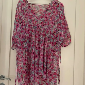 Blommig strandtunika i rosa toner - En luftig strandtunika med blommigt mönster i rosa, lila och röda nyanser. Tunikan har v-ringning, halvlånga ärmar och är tillverkad i ett tunt, transparent material som passar perfekt över baddräkten på stranden. Knytband i midjan för justerbar passform. Längd 79 cm.