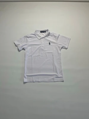 Vit pikétröja från Ralph Lauren storlek M - Klassisk vit pikétröja från Ralph Lauren med korta ärmar och krage. Tröjan har två knappar framtill och den ikoniska loggan broderad på bröstet. Perfekt basic-plagg med snygg passform och mjukt bomullsmaterial. Tröjan har storleken Medium och den är helt ny och aldrig buren