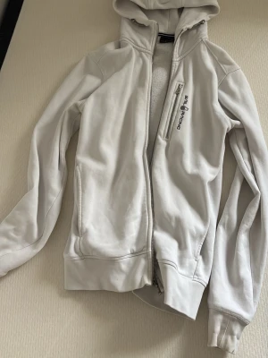 Vit hoodie från Sail Racing - Snygg vit hoodie från Sail Racing med dragkedja och huva. Har en bröstficka med dragkedja och Sail Racing-logga på bröstet. Tillverkad i mjukt bomullsmaterial med fleecefoder på insidan för extra komfort. Perfekt för en avslappnad och sportig stil. OPS! Har lite defekter på ärmmuddarna men syns knappt, skriv för fler bilder.