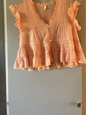 Korallfärgad volangblus med spets - Söt persikofärgad blus med volanger och spetsdetaljer. Blusen har v-ringning, knappar framtill och korta volangärmar. Lätt och luftig modell med rynkade partier och dekorativa sömmar. Perfekt för en trendig och feminin look. Jättesöt inför sommaren!! Storlek M