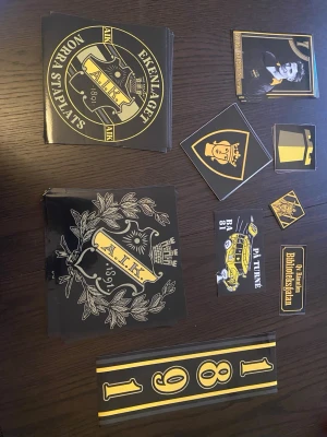AIK klistermärken samling - Samling med flera AIK-klistermärken i svart och gult. Innehåller olika motiv som klubbmärken, text som 'Ekenlaget Norra Ståplats', 'Biblioteksgatan', samt AIK:s emblem och årtal. Perfekt för att visa support för AIK på väskor, datorer eller andra ytor.