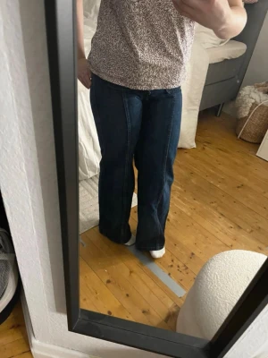 Mörkblå utsvängda jeans med fickor - Säljer ett par mörkblå jeans med hög midja och utsvängda ben. Jeansen har två stora fickor framtill med dekorativa sömmar och knappar. Klassisk denimlook som passar perfekt till en avslappnad stil. Storlek 30-31.