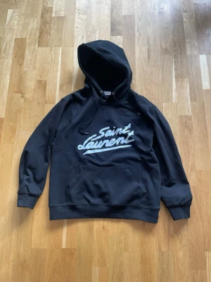 Svart hoodie från Saint Laurent - Svart hoodie från Saint Laurent med vit logga framtill i streetstil. Klassisk känguruficka och dragsko i huvan. Tillverkad i Italien, mjukt material som känns lyxigt och bekvämt. Perfekt för dig som gillar exklusiva märken och snygg design.
