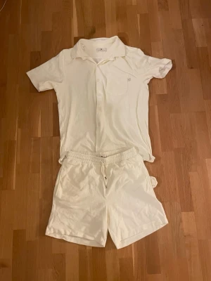 Krämvitt set med skjorta och shorts - Säljer ett krämvitt set med kortärmad skjorta och matchande shorts. Skjortan har klassisk krage, bröstficka och knappar framtill. Shortsen har resår i midjan och dragsko för skön passform. Båda delar i mjukt, tunt tyg som känns luftigt.