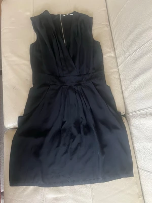 Svart omlottklänning i silkeslook - Snygg svart kortklänning med omlottliv och ärmlös design. Klänningen har markerad midja och lätt glansigt material som ger en elegant känsla. Perfekt för dig som gillar stilrena och tidlösa plagg.