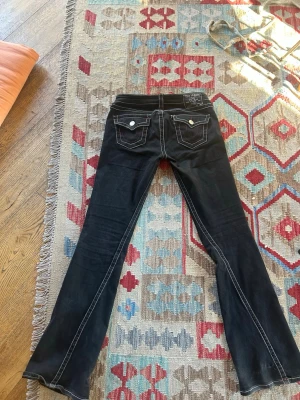 Jättefina svarta jeans från true religion - Säljer ett par jättefina svarta true religion jeans med vita och röda sömmar som tyvärr inte passar längre!!  De är lite sönder längst ner och mellan benen (se näst sista och sista bilden) men annars i väldigt bra skick.  Mått:  Rakt över midjan: 38cm Innerbenslängd: 81cm Längd: 101cm Skrev: 18cm