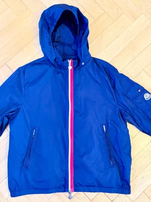 Mörkblå vindjacka med huva från Moncler - Snygg mörkblå vindjacka från Moncler med huva och dragkedja i vitt och rött. Jackan har två framfickor med dragkedja och en ficka med knapp på ärmen där Moncler-loggan sitter. Tillverkad i lätt och tåligt material, perfekt för blåsiga dagar.