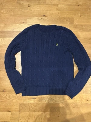 Stickad tröja Polo Ralph Lauren - Kabelstickad mörkblå tröja från Polo Ralph Lauren med rund halsringning och klassisk gul broderad logga på bröstet. Tröjan har långa ärmar och ribbade muddar vid ärmslut och nederkant. Perfekt för lager på lager och stilren look. Lappen är av!