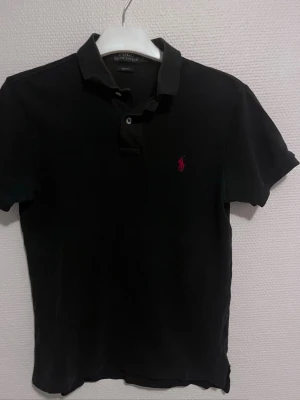 Svart Polo Ralph Lauren pike S - Svart pike från Polo Ralph Lauren i slim fit . Pike. är gjord i 100% mjuk bomull. På bröstet finns den ikoniska röda broderade Ralph Lauren loggan.