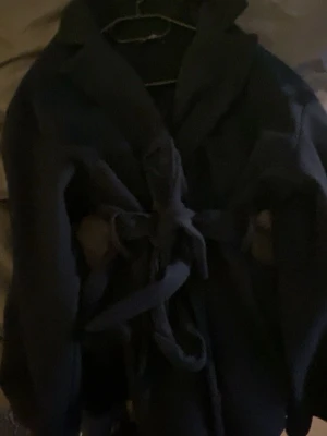 Marinblå kappa med knytskärp - Stilren marinblå kappa med klassisk krage och knytskärp i midjan. Kappan har en rak passform och är perfekt för kyligare dagar. Materialet ser ut att vara ullblandning och modellen är tidlös och enkel att matcha.