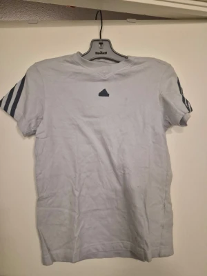 Vit t-shirt med blå ränder Adidas - Vit t-shirt från Adidas med blå ränder på ärmarna och över ryggen. Klassisk Adidas-logga på bröstet. T-shirten har rund hals och är kortärmad, perfekt för en sportig och avslappnad stil.