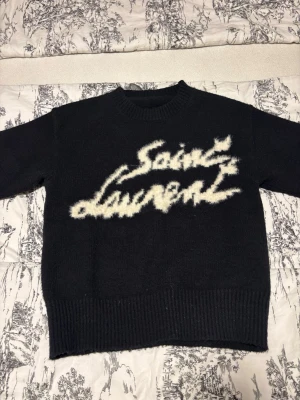 Svart stickad Saint Laurent-tröja - Stickad svart tröja från Saint Laurent med vit fuzzy logotyp framtill. Tröjan har rund hals, långa ärmar och ribbstickade muddar vid ärmslut och nederkant. Perfekt för dig som gillar streetstyle och exklusiva märken.