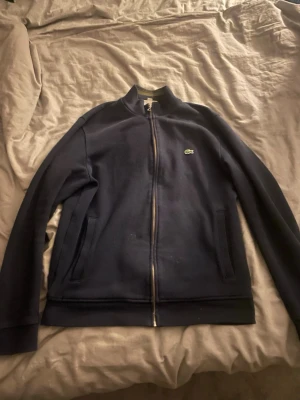 Mörkblå zip hoodie från Lacoste - Snygg mörkblå hoodie från Lacoste med dragkedja framtill och två fickor. Klassisk krokodillogga på bröstet och ribbade muddar vid ärmar och nederkant. Tillverkad i mjukt bomullsmaterial för en bekväm känsla. Perfekt för en avslappnad och stilren look.