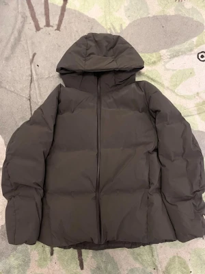 Svart pufferjacka med huva - En grå pufferjacka med huva och dragkedja framtill. Jackan har en boxig passform och breda kanaler som ger en modern look. Perfekt för kalla dagar när du vill hålla värmen och ändå se snygg ut. Materialet är slätt och jackan är vadderad för extra värme.