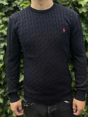 Svart kabelstickad tröja Polo Ralph Lauren - Kabelstickad svart tröja från Polo Ralph Lauren med rund halsringning och ribbade muddar. Tröjan har det klassiska röda Polo-logotypen broderad på bröstet och är tillverkad i ett mjukt material. Perfekt för en stilren och avslappnad look.