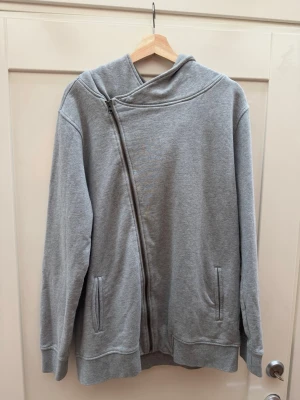 Grå hoodie med sned dragkedja A Mid.Munk - Snygg grå hoodie med unik sned dragkedja framtill och två fickor. Hoodien har en stor huva och är tillverkad i mjukt sweatshirtmaterial. Perfekt för en chill och avslappnad stil. A Mid.Munk product for the congregation