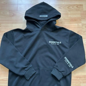 Svart Essentials Fear of God hoodie - Svart hoodie från Essentials Fear of God med vit logga tryckt på bröstet och ärmen. Klassisk oversized passform, stor huva och känguruficka framtill. Tillverkad i mjukt bomullsmaterial som är perfekt för en avslappnad streetwear-look.
