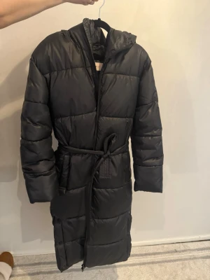 Svart lång dunjacka med huva - Säljer en svart lång dunjacka med bälte i midjan och huva. Jackan har dragkedja framtill och stora, quiltade partier som ger en riktigt puffig look. Perfekt för kalla vinterdagar och riktigt skön att mysa in sig i.