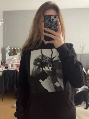 Svart Tupac hoodie med tryck - Snygg svart hoodie med stort svartvitt tryck av Tupac på bröstet. Klassisk huva med dragsko och avslappnad passform. Perfekt för dig som gillar streetwear och vill ha en hoodie med attityd.