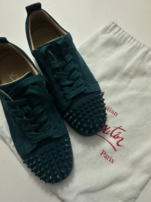 Mörkgröna sneakers från Christian Louboutin - Säljer ett par mörkgröna sneakers från Christian Louboutin i mocka med matchande gröna skosnören. Skorna har coola nitar på tån och klassisk låg silhuett. Perfekta för dig som vill sticka ut med en edgy och lyxig stil. Skorna är använda 2-3 gånger och är som nya, tyvärr har jag fått fläckar på undersidan därav det billiga priset! 