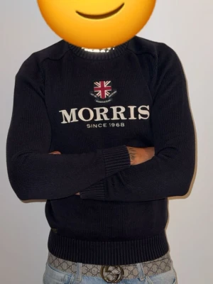 Svart tröja från Morris - Snygg svart stickad tröja från Morris med broderad logga och brittisk flagga på bröstet. Rund halsringning och ribbade muddar vid ärmslut och nederkant. Perfekt för en stilren och klassisk look.