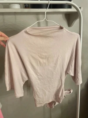 Ljusrosa topp med knyt i midjan - Trendig ljusrosa topp med trekvartsärm och knytband i midjan. Tillverkad i mjukt, tunt bomullsliknande material och har en enkel, avslappnad passform. Perfekt att styla med höga jeans eller kjol för en cool look.