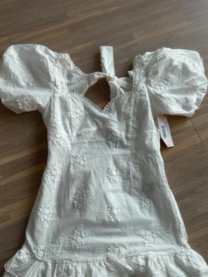 Studentklänning  - Supersöt vit kort klänning med puffärmar, broderade blommor och volangkant. Klänningen har en öppen rygg med knytband och v-ringad hals. Perfekt för sommarens alla tillfällen och ger en romantisk vibe.