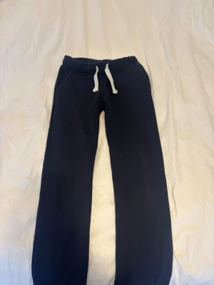 Mörkblå mjukisbyxor från Russemerch - Mörkblå mjukisbyxor från Russemerch med vita dragsnören i midjan och två sidofickor. Byxorna har en klassisk rak passform och är gjorda i mjukt, fleeceliknande material som är riktigt skönt mot huden. Perfekta för chill dagar eller när du vill vara extra bekväm.