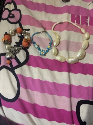 Mix av armband och halsband i färg - Säljer ett set med tre smycken: ett chunky armband med stora pärlor och orange detaljer, ett blått armband med blommor och pärlor samt ett halsband med vita snäckformade pärlor. Perfekt för dig som gillar färgglada och unika accessoarer. Alla de tre tillsammans kostar 20 kronor men för en kostar det 10 kr 