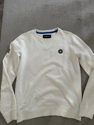 Vit sweatshirt Sail Racing - Vit sweatshirt från Sail Racing  med rund halsringning och broderad logga på bröstet. På ryggen står det 'USA 77' i svart text och på ena ärmen finns en amerikansk flagga. Tröjan har ribbade muddar och är tillverkad i mjukt bomullsmaterial.