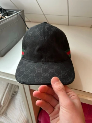 Svart Gucci keps med GG-mönster - Säljer en svart keps från Gucci med klassiskt GG-monogram över hela kepsen. På sidan finns en grön och röd rand som ger den en ikonisk look. Justerbar baktill för perfekt passform. Materialet är troligtvis bomullscanvas. Går att pruta. Äkta! 