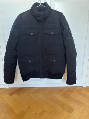 Svart Woolrich jacka - Säljer en svart pufferjacka med fyra framfickor med lock och knappstängning. Jackan har hög krage, ribbade muddar och en klassisk quiltad design. Perfekt för gråa dagar och har en stilren look som passar till det mesta. Skick 5/5, strlk US: M, EU: L. Nypris: 6000