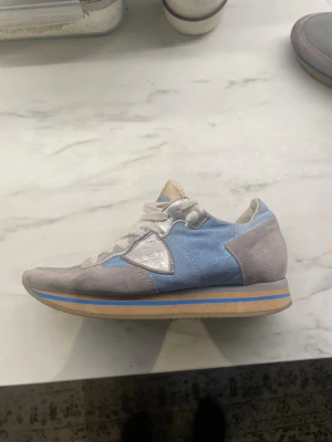 Blå och grå sneakers från Philippe Model - Snygga sneakers från Philippe Model i ljusblått tyg och grå mocka med vita detaljer. Skon har en klassisk låg siluett, vita skosnören och en beige sula med blå rand. Perfekt för dig som gillar stilrena och sportiga sneakers.