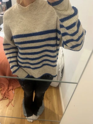 Beige stickad tröja med blå ränder - Säljer en oversized beige stickad tröja med blå horisontella ränder. Tröjan har hög hals, långa ärmar och slitsar i sidorna. 