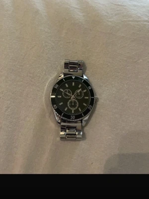 Stilren silverfärgad klocka - Snygg klocka med metallarmband i silver och svart urtavla med tre mindre visartavlor. Klassisk rund form och markerade siffror runt boetten. Perfekt accessoar för dig som gillar en clean och tidlös look.