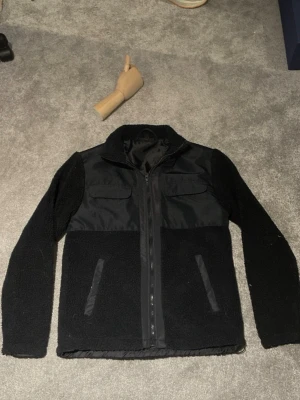 Svart fleecejacka från Saligia XS - Säljer en svart cardiganjacka från Saligia i storlek XS. Jackan har en mix av fleece och slätare tyg, två bröstfickor med lock och två snedställda fickor framtill. Dragkedja hela vägen och hög krage för extra värme.