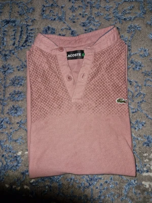Rosa Lacoste t-shirt med mönster - Säljer en rosa t-shirt från Lacoste med korta ärmar och diskret mönster upptill. Tröjan har knappar vid halsen och den klassiska Lacoste-loggan på bröstet. Perfekt för en stilren och avslappnad look.