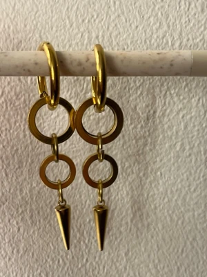 Guldiga örhängen  - Säljer ett par statement örhängen i guldfärgad metall. De har stora hoops med två mindre ringar som hänger ner och avslutas med en spetsig kon. Snyggt och edgy design som verkligen sticker ut. Perfekta för dig som gillar unika accessoarer.