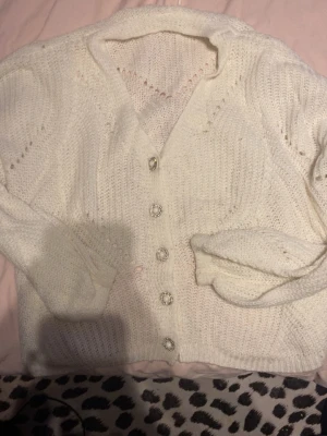 Vit stickad kofta med jätte fina knappar - Säljer en söt vit stickad kofta med långa ärmar och v-ringning. Koftan har dekorativa pärlknappar med glittrande detaljer och ett mjukt, luftigt material. Perfekt att slänga över en topp för en mysig look.